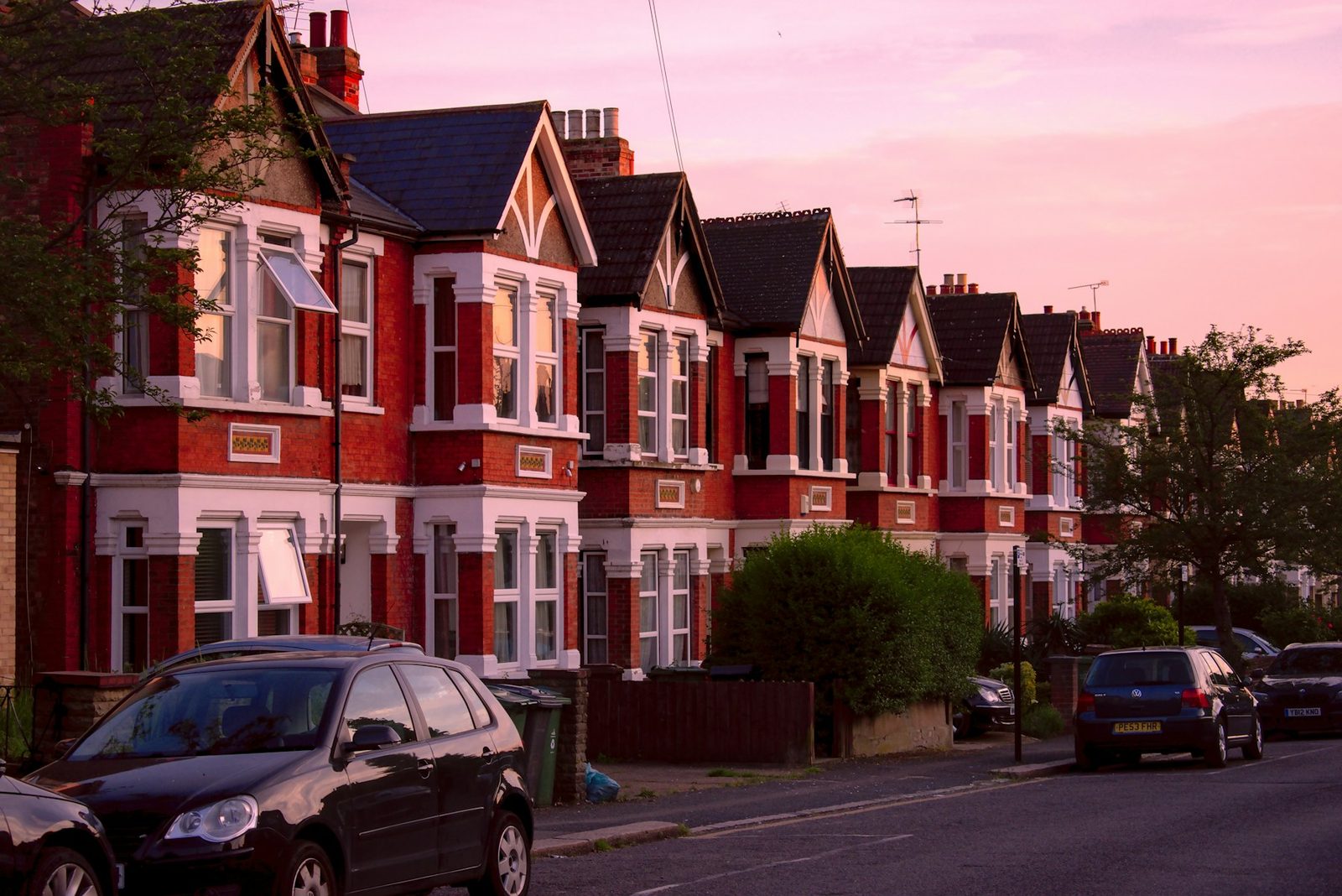 A row of Victorian brick terraced houses on a residential street in London, bay windows and low front walls, overcast daylight.