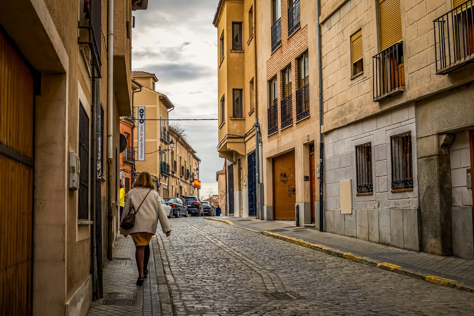 A quiet cobblestone street in the old quarter of a Spanish town, stone and painted buildings, a lone pedestrian, flat daytime light.