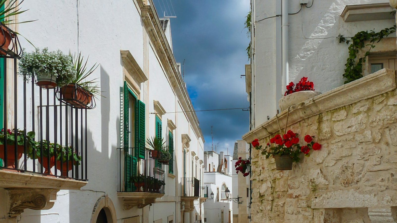 A narrow whitewashed street in the old town of Ostuni, in Puglia, southern Italy, green shutters and pots of red geraniums along the walls.