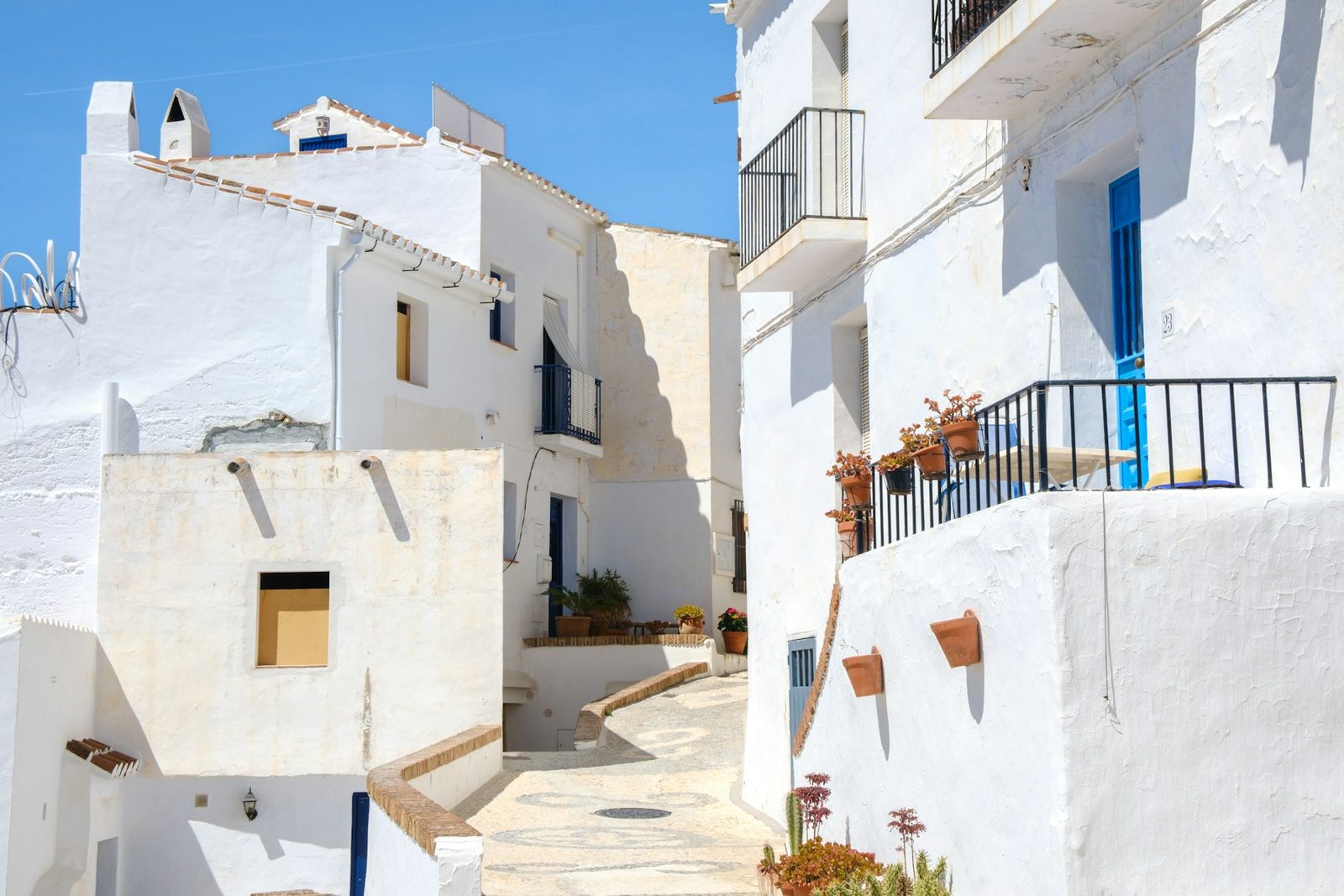 A narrow sunlit street between traditional whitewashed buildings in a Spanish village.